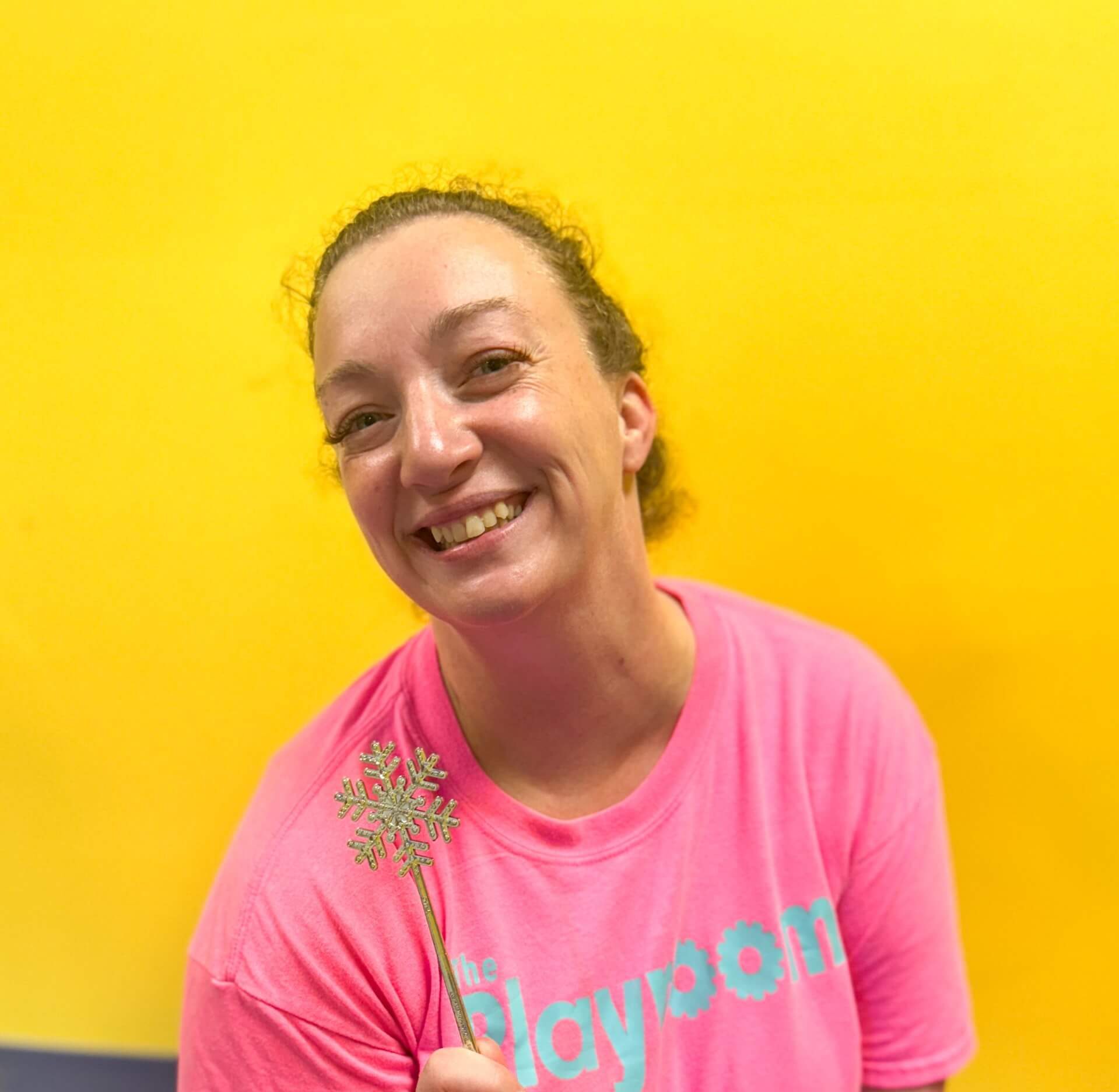 Katie Hay sits on a the floor in The Playroom dance and activity room with yellow walls. She is holding a magic wand and smiling wearing a neon pink Playroom tshirt