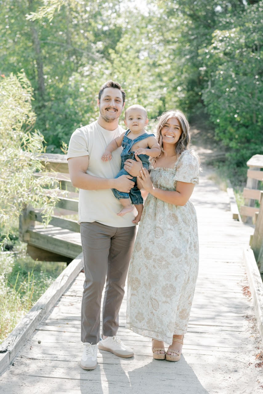Miss Mel, Pre-K 3 teacher at The Playroom, stands with her husband and baby for family portrait in outdoor setting
